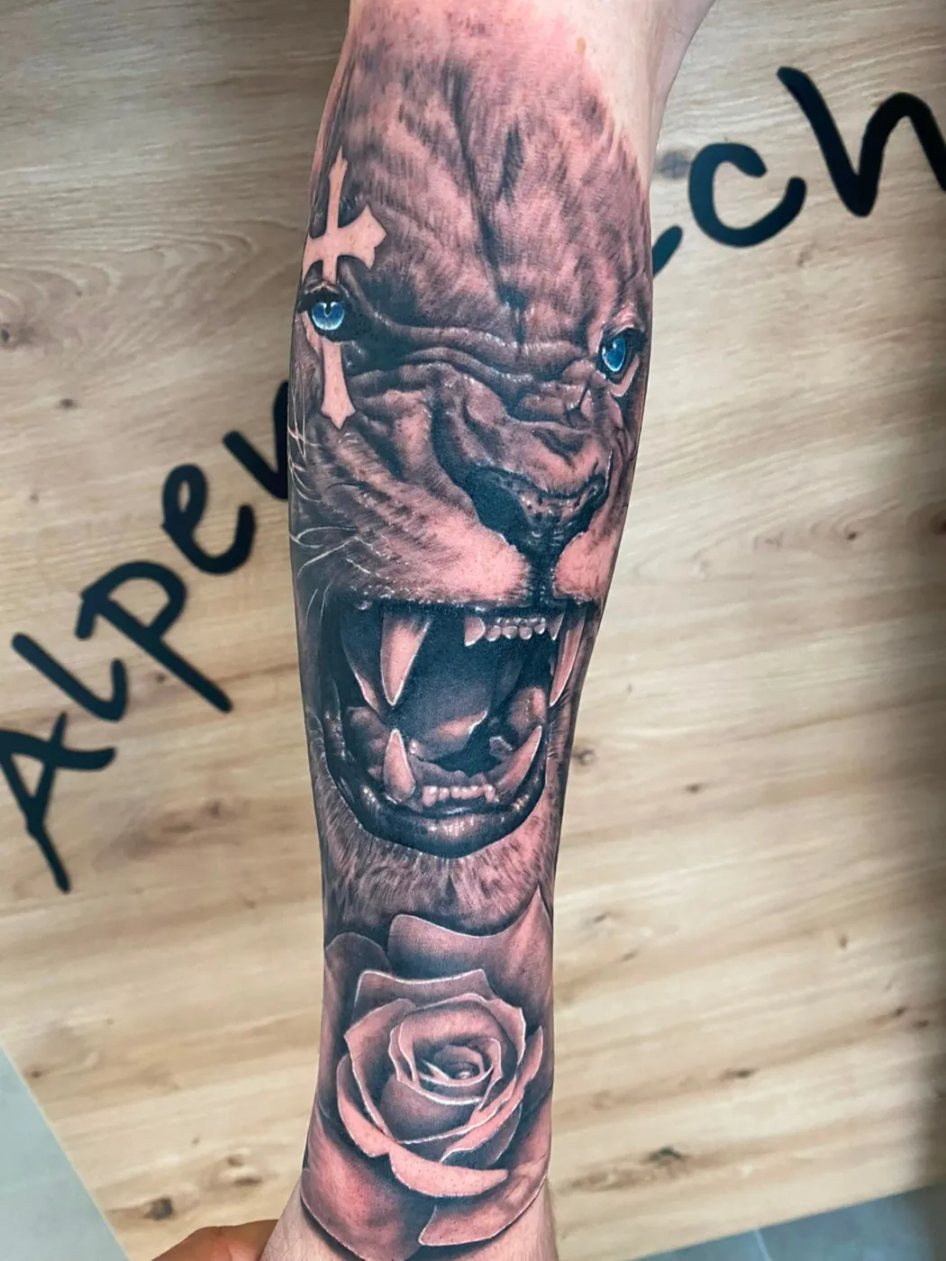 Realistischer brüllender Löwe mit blauen Augen, Kreuz und Rose am Unterarm — tätowiert von Han, Alpenstich Windischgarsten